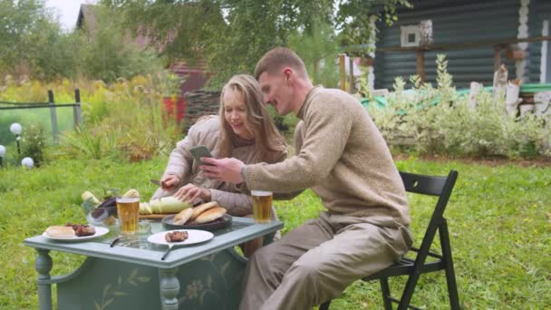 Een jonge blonde vrouw snijdt een courgette in de achtertuin van een dacha, een man toont haar grappige foto 's op zijn smartphone — Stockvideo