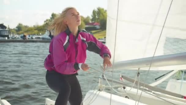 美しい若い女性は、晴れた夏の日にレガッタを航海する川のためにヨットを準備します。帆を上げるためにロープを引く. — ストック動画