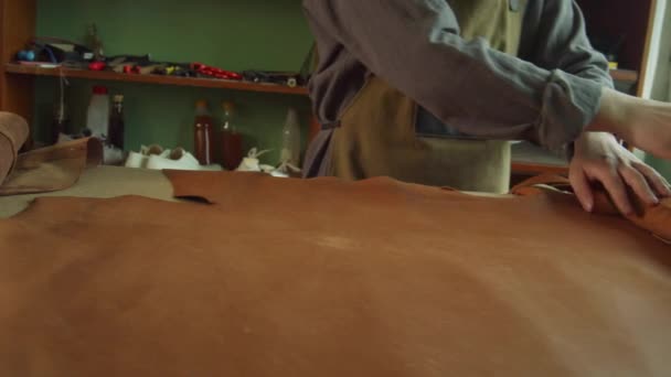 En erfaren skräddare rullar ut en rulle äkta läder på arbetsbänken. Förberedelse för produktion av en exklusiv väska för kvinnor. Långsam. — Stockvideo