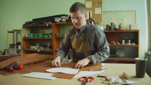 Un esperto Tanner maschio realizza un layout di elementi della borsa su un grande pezzo di pelle marrone su un banco da lavoro in officina. — Video Stock