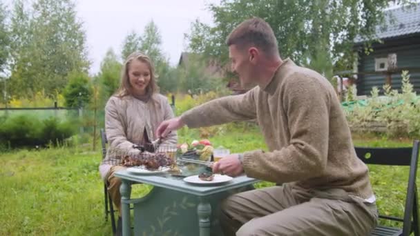 Муж ухаживает за женой за столом, кладет мясо на тарелку, помещает в карантин в деревне — стоковое видео