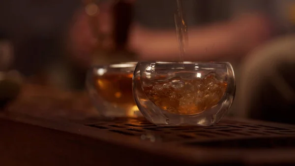 Cozy atmosphere of a tea room in a Chinese province. A stream of fragrant tea from a porcelain teapot fills two glass bowls. Close-up, warm light. Slowmo. — Stock Photo, Image