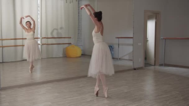 Seorang balerina dalam pakaian balet putih melakukan berbagai gerakan balet di depan cermin di ruang dansa selama latihan — Stok Video