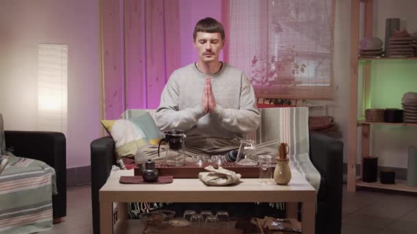 Een man in een grijze trui bidt met gevouwen handen tijdens een theeceremonie voordat hij thuis thee drinkt — Stockvideo