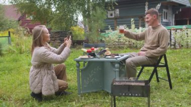 Genç bir adam birayla fotoğraf çektirmek için poz veriyor. Bir kız akıllı telefondan onun fotoğraflarını çekiyor. Köy evinin arka planıyla karşılaştırıyor..