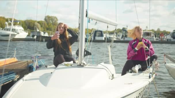 Dwie siostry siedzące na pokładzie białego jachtu sportowego robią sobie selfie na smartfonach i relaksują się po wyścigu żeglarskim. — Wideo stockowe