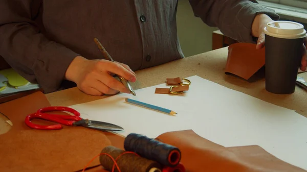 Un tailleur expérimenté dans l'atelier derrière l'établi prépare des accessoires pour la création d'un sac en cuir. Colle ensemble des éléments et boit du café pendant la pause. — Photo