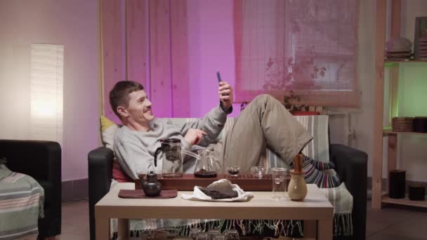 Un giovane uomo con i baffi è sdraiato su un divano in una stanza sull'auto-isolamento e comunica con gli amici tramite video chat. Davanti a lui sul tavolo c'è un set per la cerimonia del tè cinese. — Video Stock