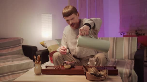 Ein Mann sitzt zu Hause auf einem Sofa und gießt kochendes Wasser aus einer Thermoskanne in eine Teekanne für eine Teezeremonie — Stockvideo