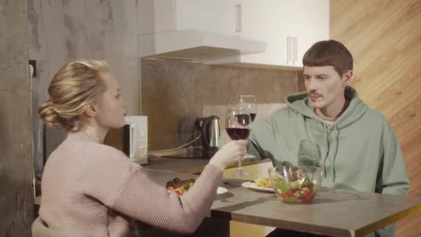 Os jovens, um homem e uma mulher, bebem vinho tinto durante um jantar festivo, comem massa e colam copos. Interior de uma cozinha acolhedora. — Vídeo de Stock