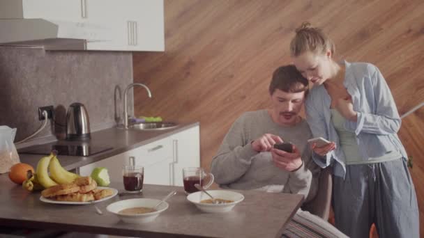 Mąż dzieli się z żoną sukcesem w ich pracy. Pokazuje coś interesującego na ekranie smartfona. Dziewczyna się uśmiecha i jest szczęśliwa. Kuchnia wnętrza. — Wideo stockowe