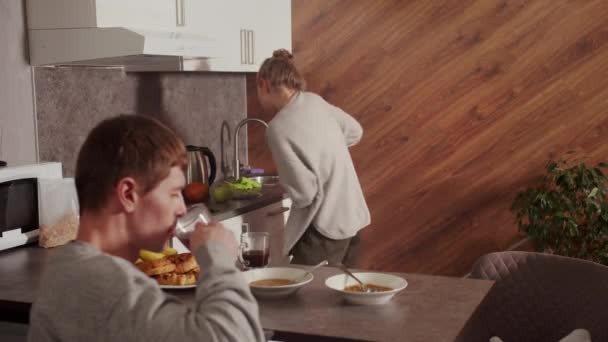 Uma típica manhã de família, a esposa está ao telefone e limpando a cozinha, o marido olha para ela e bebe chá — Vídeo de Stock