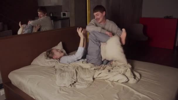 Linda chica y su marido juegan en la cama, pelean con almohadas, se divierten juntos de vacaciones en un hotel, disfrutan el uno del otro — Vídeo de stock