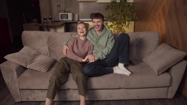 Una alegre pareja joven ver una divertida película Comedia en la televisión, sentado en un sofá acogedor. Se acurrucan y se divierten por la noche.. — Vídeo de stock