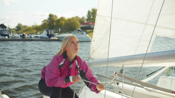 A beautiful young woman prepares her yachts rigging for a river sailing regatta on a sunny summer day. Pulls the rope to raise the sail. — Stock Photo, Image