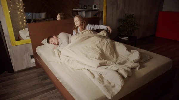 O fată în pijamale merge la culcare cu soțul ei adormit și îmbrățișează ușor bărbatul sub pătură. Lumina soarelui dimineața devreme în interiorul dormitorului . — Fotografie, imagine de stoc
