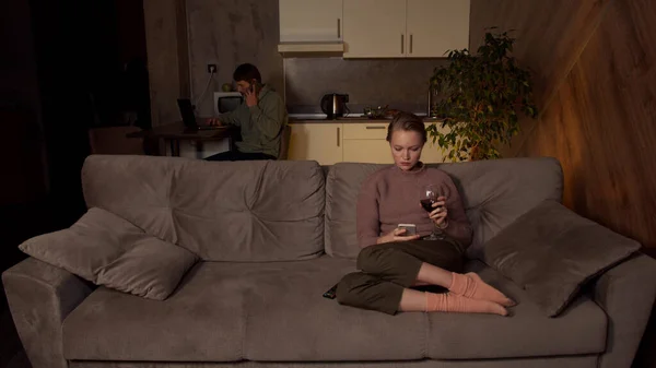 Una chica con ropa acogedora está sentada en el sofá de la sala de estar, bebiendo vino y viendo la televisión. Su marido está en la mesa de la cocina, trabajando en un ordenador portátil, haciendo llamadas de trabajo en un teléfono inteligente. — Foto de Stock