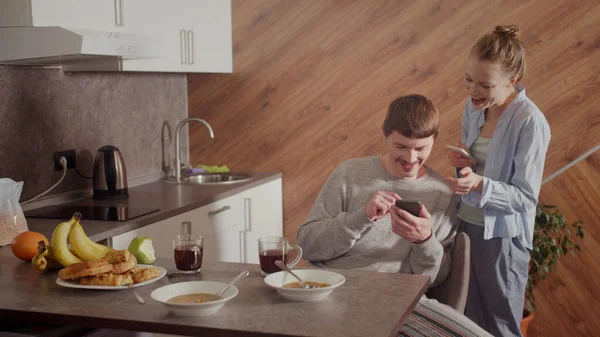 El marido comparte con su esposa el éxito en su trabajo. Muestra algo interesante en la pantalla del teléfono inteligente. La chica sonríe y es feliz. Interior de cocina. — Foto de Stock