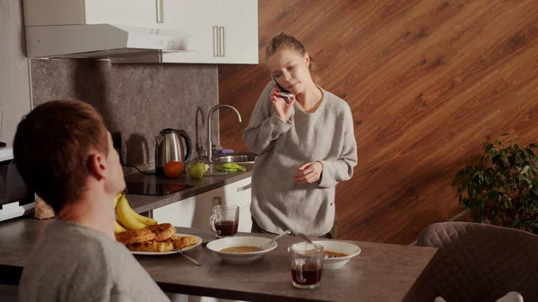 Una típica mañana familiar, la esposa está al teléfono y limpiando la cocina, el marido la mira y bebe té — Foto de Stock