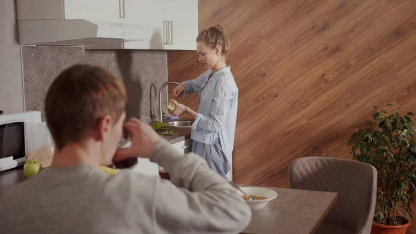 Una joven está fregando una manzana sobre el fregadero y hablando con su novio en la cocina — Foto de Stock