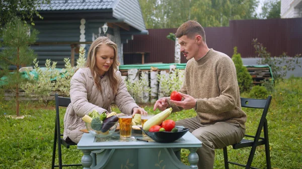 Mientras se fríe la barbacoa, una mujer corta verduras en una mesa en el patio trasero de la casa y habla con un hombre. — Foto de Stock