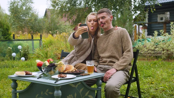 Una pareja joven toma una selfie en una mesa de picnic en el país en un teléfono inteligente, muestra el idioma — Foto de Stock