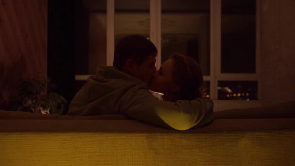Przytulny wieczór faceta i dziewczyny. Małżeństwo całuje się w usta, siedzi na kanapie w pokoju na tle świateł nocnego miasta. — Wideo stockowe