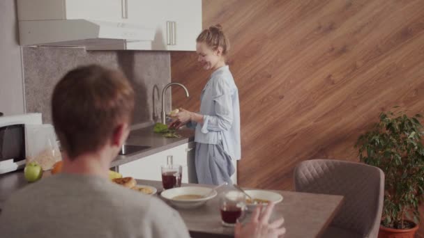 Вранці, на кухні, хлопець снідав за столом, дівчина знімає шкірку з яблука і їсть її — стокове відео