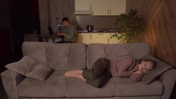 Un uomo con uno smartphone sta lavorando su un computer portatile in cucina mentre sua moglie dorme sul divano in soggiorno. — Video Stock