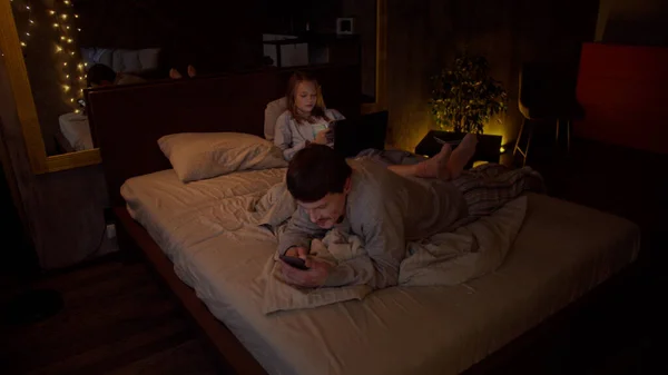 Egy fiatal pár és a barátja az ágyon fekszenek, telefonnal a kezükben, beszélgetnek, a lánynak laptopja van. — Stock Fotó