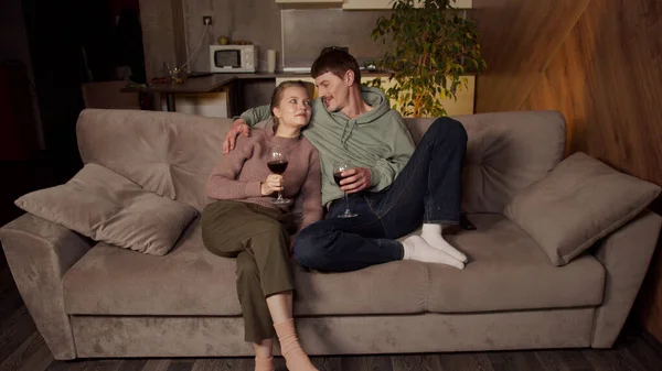 Acogedora velada familiar con una copa de vino. Un chico y una chica se relajan en el sofá, viendo una película. Vista desde el televisor al apartamento estudio. — Foto de Stock