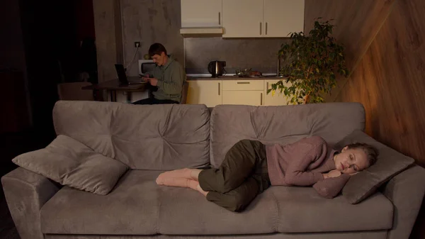 Akıllı telefonlu bir adam, karısı oturma odasındaki kanepede uyurken mutfakta dizüstü bilgisayarla çalışıyor..