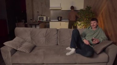 Kadın bir aile yemeğinden sonra mutfağı temizlerken, koca bir bardak şarapla televizyon izliyor. Aile ve cinsiyet eşitsizliği kavramı..