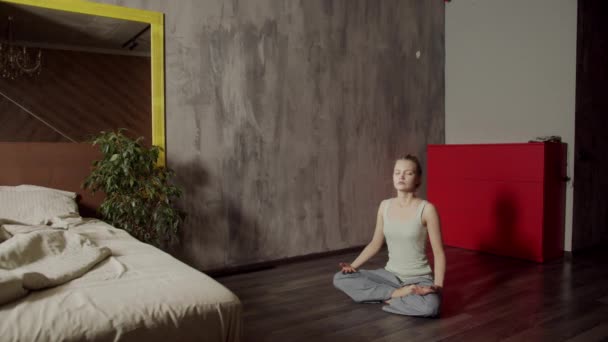 Genç bir kız Budist yogasının temellerini evde öğreniyor. Nefes alma ve omurga sağlığını eğitmek için basit asmalar yapar. Lotus asana. — Stok video