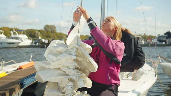 A young blonde with a teammate puts the mainsail on the afterstay of her sports yacht. Straightens the sails and prepares the rigging for the race. — Stock Photo, Image