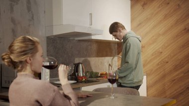 Akşamları, mutfakta, bir adam salata için sebze keserken, misafir kızı mutfak masasında oturmuş kırmızı şarap içiyor.