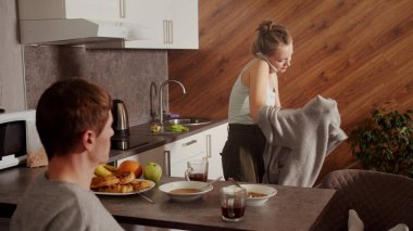 Koca sabah sakince çay içerken, kadın işe geç kalıyor, telefonda konuşuyor ve çabucak üstünü değiştiriyor.