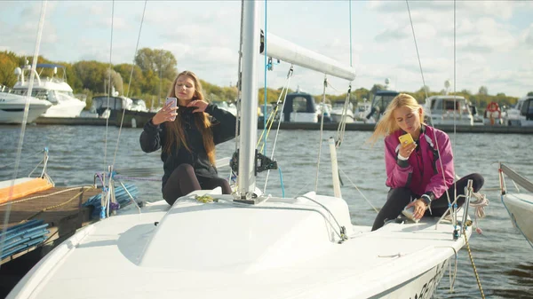 Две сестры, сидящие на борту белой спортивной яхты, делают селфи на смартфонах и отдыхают после парусной гонки.. — стоковое фото
