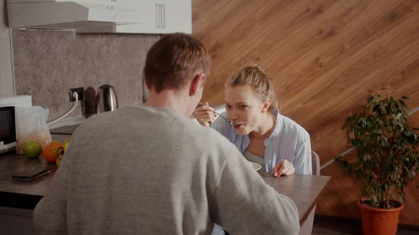 La chica le pasa a su marido una bolsa de copos de maíz para el desayuno. Desayunan juntos en la cocina en un día libre. — Foto de Stock