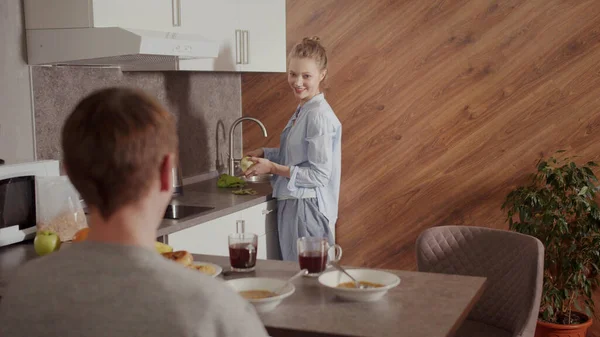 Eine glückliche junge Frau bereitet einem Mann ein gesundes Frühstück zu. Steht an der Spüle, schält einen grünen Apfel und spricht mit ihrem Mann. — Stockfoto