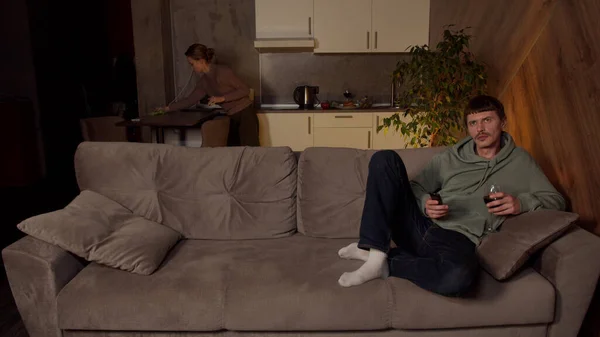 Kadın bir aile yemeğinden sonra mutfağı temizlerken, koca bir bardak şarapla televizyon izliyor. Aile ve cinsiyet eşitsizliği kavramı..