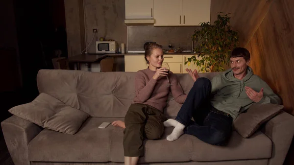 Joven adulto chico y chica están sentados en el sofá de un acogedor apartamento, charlando y riendo. Un hombre le da a una mujer una copa de vino tinto para tomar un sorbo.. — Foto de Stock