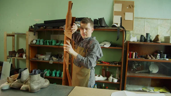 Молодой обувной мастер берет рулон натуральной кожи с полки и кладет его на рабочее место, готовясь к работе. — стоковое фото