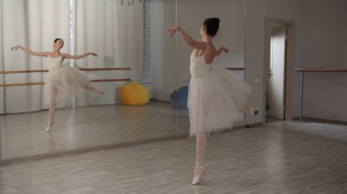 Beyaz bale kıyafeti giymiş bir balerin prova sırasında dans salonundaki aynanın önünde çeşitli bale hareketleri yapar.
