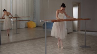 Güzel bir balerin bale korkuluğunun önünde duruyor ve bacaklarını aynanın önünde açıyor.