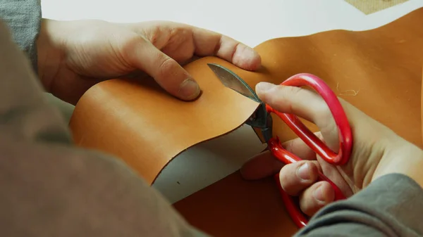 De handen van een kleermaker maken een precieze en gelijkmatige snede van een stuk leer met een schaar voor het maken van een handgemaakt product te koop. — Stockfoto