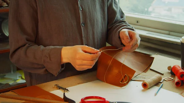 Un jeune apprenti tailleur expérimenté enfile une aiguille pour coudre un produit en cuir véritable dans un atelier de cuir. — Photo