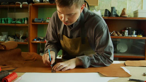 Un jeune tanneur fait des marques avec un crayon et une règle sur un morceau de cuir véritable pour faire des trous pour les attaches sur un sac en cuir. — Photo