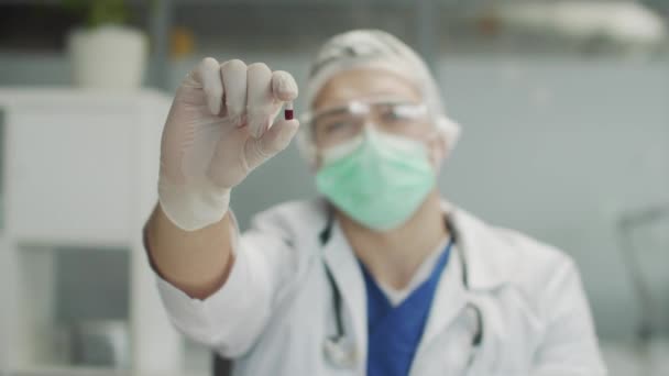 Az orvos a gyógyszeres kapszulát mutatja a kamerának. Közelkép homályos háttérrel. Az orvos védőszemüveget, kesztyűt és maszkot visel, lassított mozgás.. — Stock videók