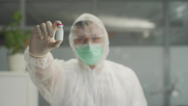 Un medico irriconoscibile in una tuta protettiva batteriologica tiene in mano un vaccino da primo piano e annuisce alla testa — Video Stock